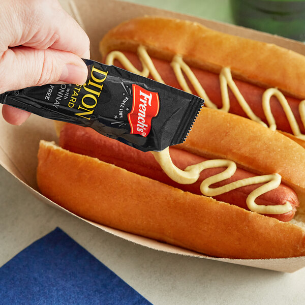 A hand holding a French's Dijon Mustard packet over two hot dogs.