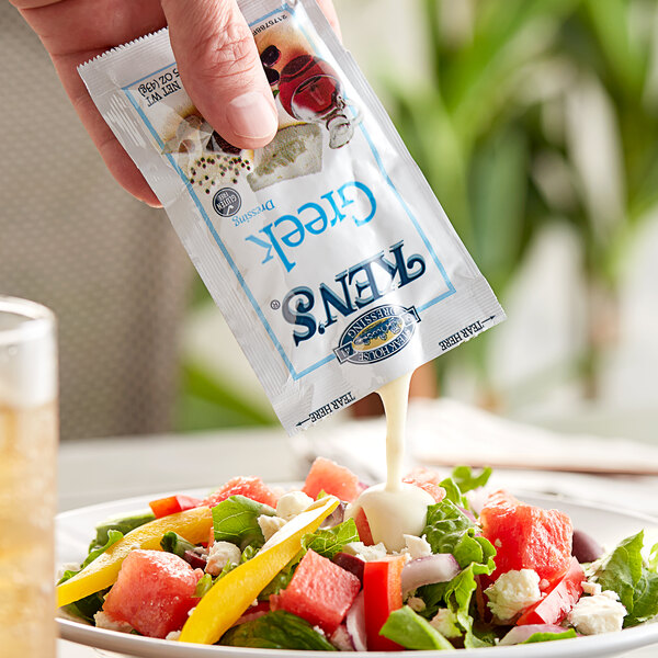 A hand pouring a Ken's Foods Greek Dressing packet over a salad.