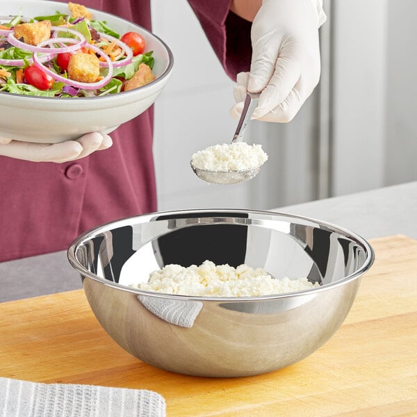 A person in a white coat holding a stainless steel mixing bowl of salad with croutons and onions.
