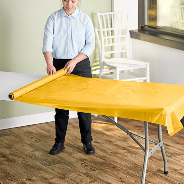A person rolling a Choice Harvest Yellow plastic table cover on a table.