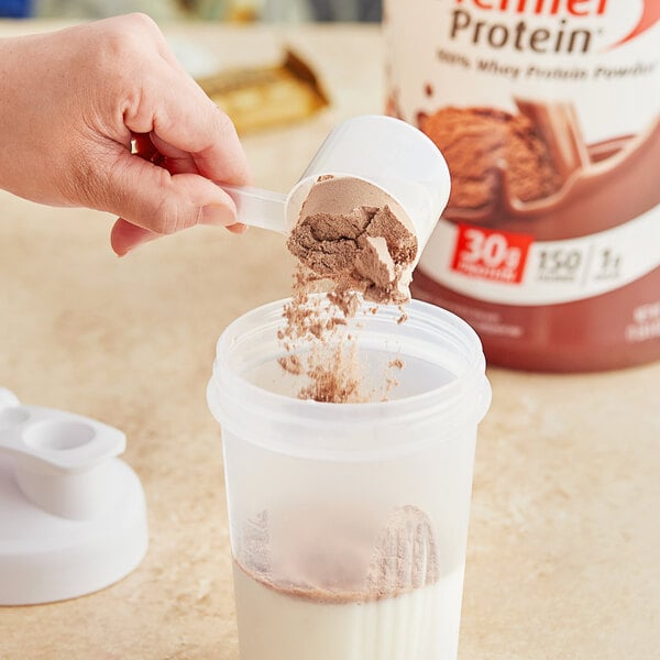A hand pouring Premier Protein chocolate powder into a blender.