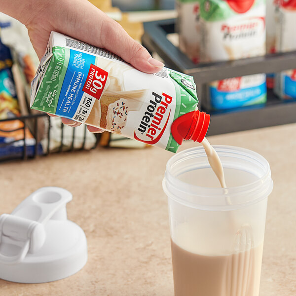 A hand pouring Premier Protein Cake Batter Delight shake from a container into a cup.