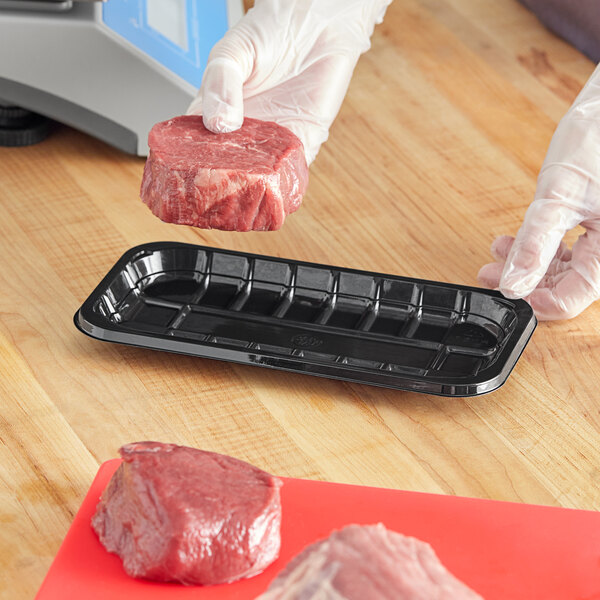 A gloved hand holding a black PET plastic meat tray on a cutting board.