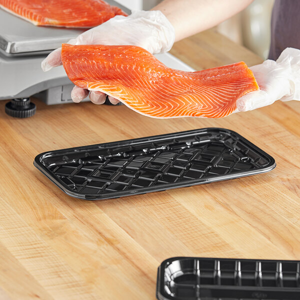 A person in gloves using a CKF black PET plastic meat tray to hold a piece of salmon on a wood surface.