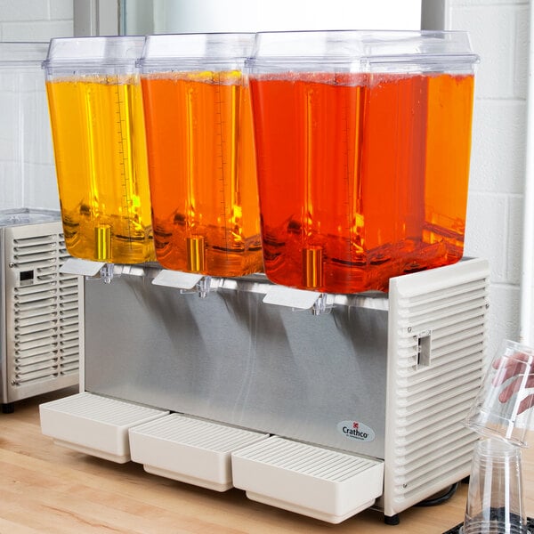 A Crathco refrigerated beverage dispenser with three clear containers of orange liquid.