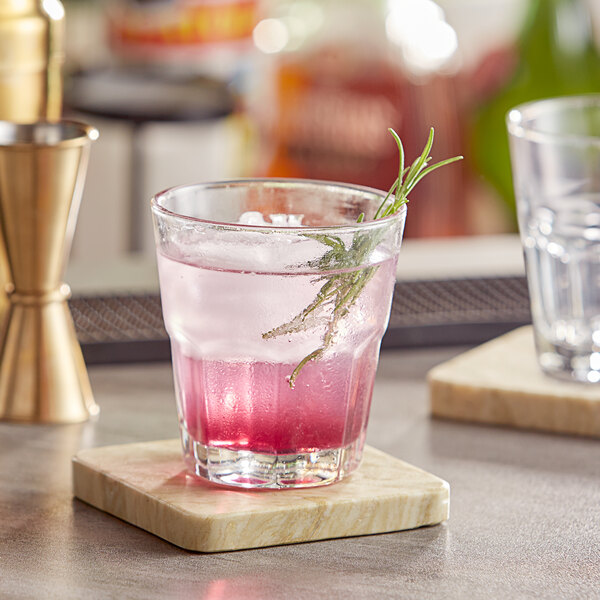 A Memphis rocks glass with pink liquid and a rosemary sprig on a wooden coaster.