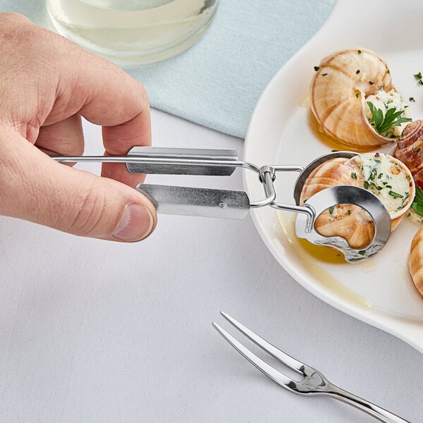 A person using Acopa stainless steel snail tongs to hold a snail shell over a plate of food.