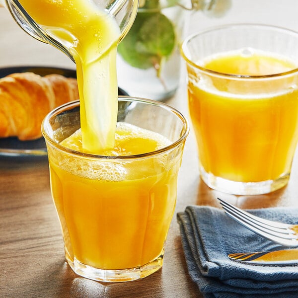A person pouring Growers' Pride Orange Juice into a glass on a table with a croissant.