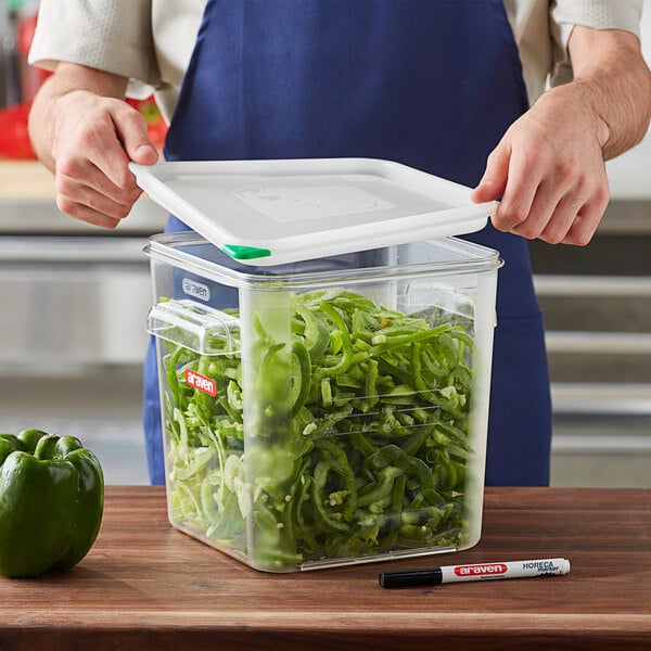 A person in a blue apron holding an Araven clear plastic container filled with green bell peppers.