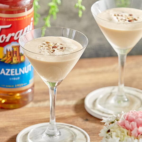 A table with two martini glasses filled with brown liquid next to a bottle of Torani Hazelnut Flavoring Syrup.