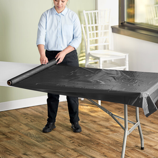 A woman rolling a Table Mate black plastic table cover on a table.