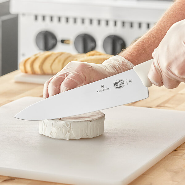 A person holding a Victorinox Chef Knife with a white handle cutting a piece of cheese.