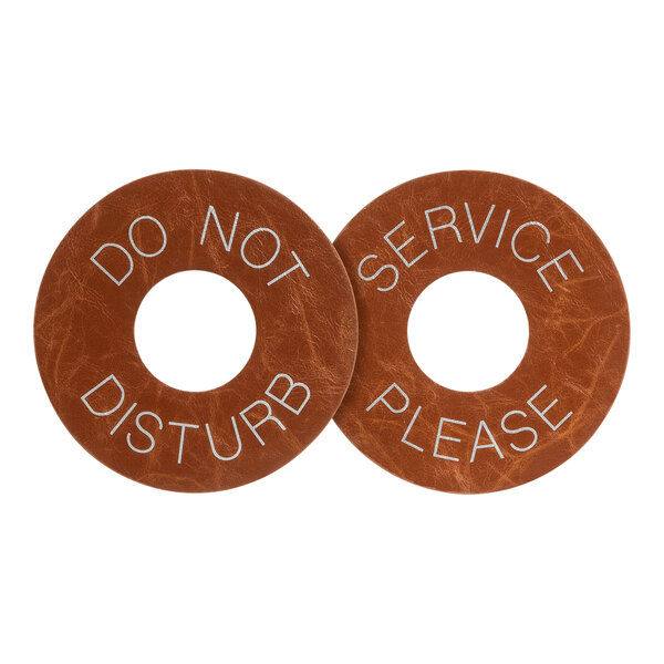 A brown faux leather round door hanger with white text reading "do not disturb" and "please service" on a table.