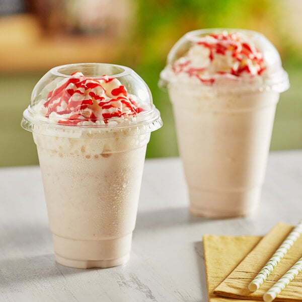 Two Choice clear plastic cups of milkshakes with whipped cream and red sprinkles with dome lids.