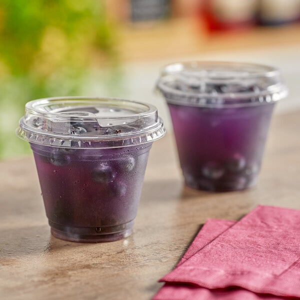Two Choice clear plastic cups of blueberry juice with lids on a counter.