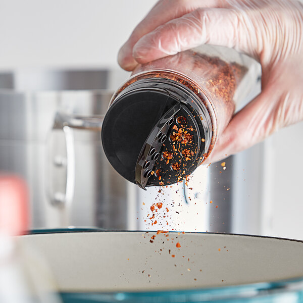 A person in gloves pouring seasoning into a container using a 63/485 black dual-flapper spice lid.