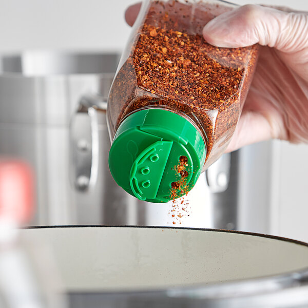 A person holding a plastic container with a green dual-flapper spice lid and pouring seasoning into it.