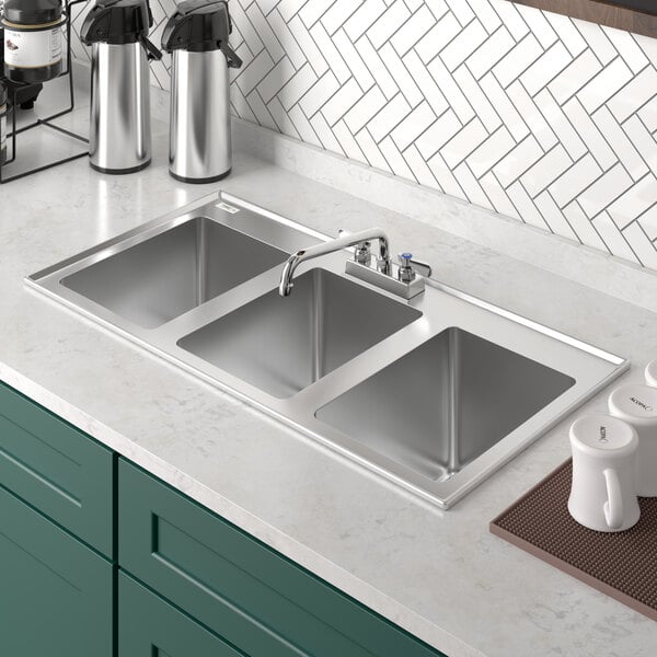 A Regency stainless steel three compartment drop-in sink with a faucet above a brown mat with a white mug.
