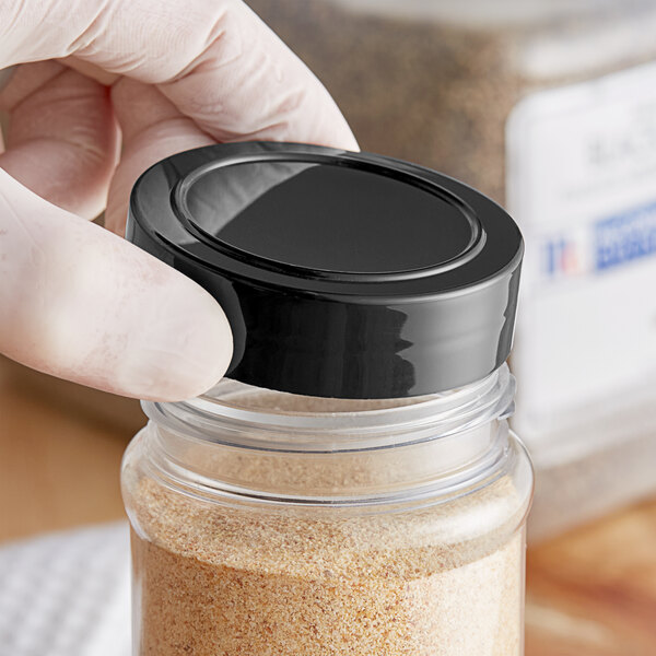 A hand holding a black 63/485 unlined polypropylene spice cap over a jar of brown powder.