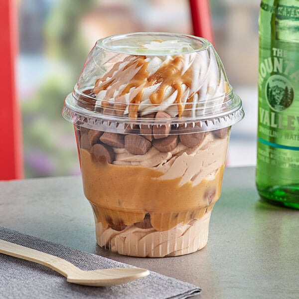 A clear plastic dessert cup filled with ice cream and caramel toppings with a low dome lid on a table.
