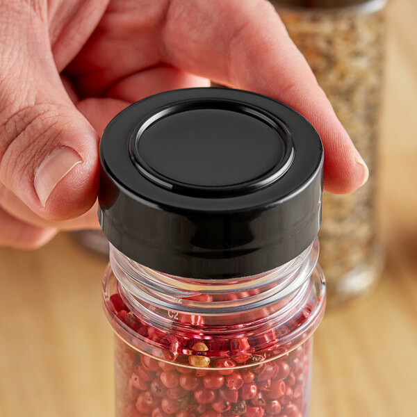 A hand holding a jar with a black unlined polypropylene spice cap filled with red and white peppercorns.