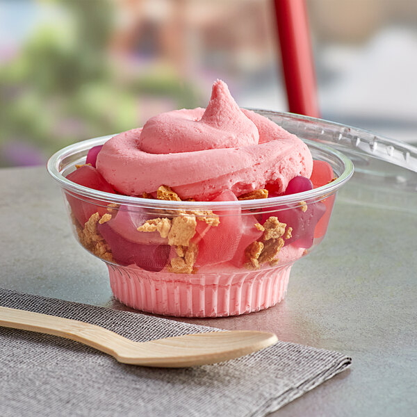 A pink bowl of ice cream with cereal and a wooden spoon on a wooden board with a clear plastic lid on the table.