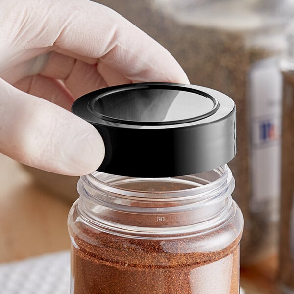 A hand holding a black 63/485 Induction-Lined Spice Cap on a jar of powder.