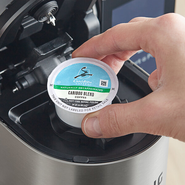 A hand placing a Caribou Coffee Decaf K-Cup pod in a coffee maker.