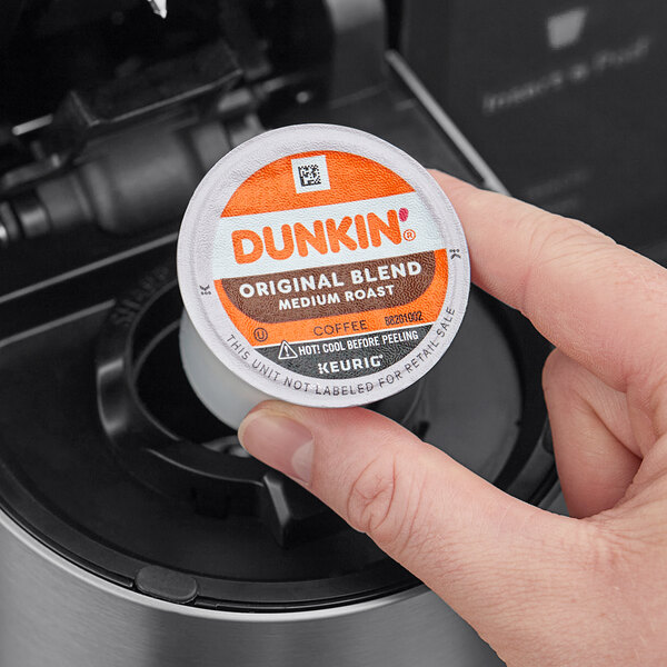 A hand holding a small round container of Dunkin' Original Blend Coffee Single Serve K-Cup Pods.