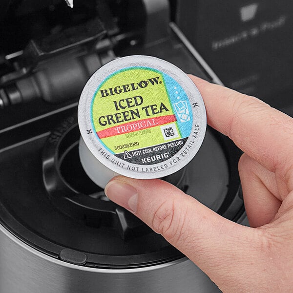 A hand holding a round container of Bigelow Iced Tropical Green Tea K-Cup Pods with a label.