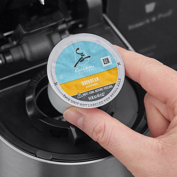 A hand holding a round container of Caribou Coffee Daybreak Morning Blend Single Serve K-Cup Pods.