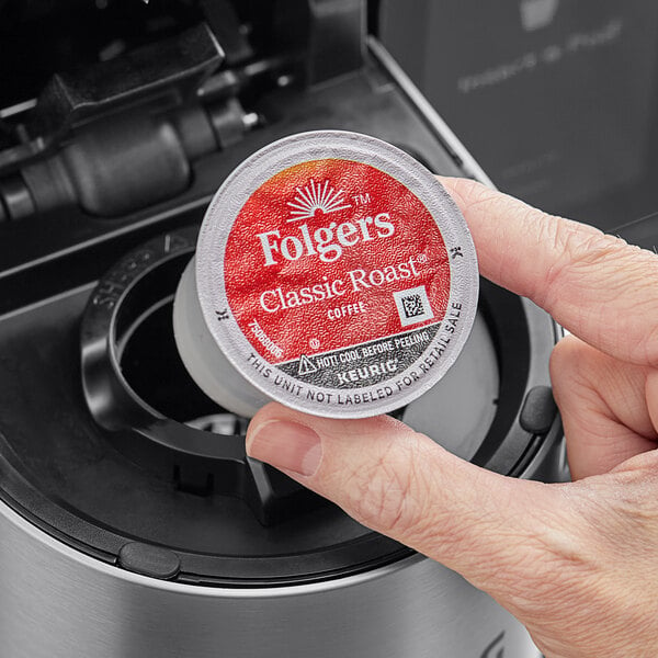A hand holding a round red and white container of Folgers Classic Roast Coffee Single Serve K-Cup Pods.