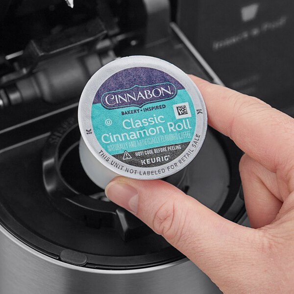 A hand holding a round blue and white container of Cinnabon Classic Cinnamon Roll coffee pods.