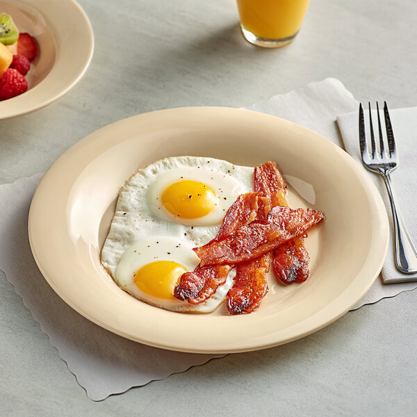 A tan Acopa Foundations wide rim melamine plate with two eggs, bacon, and fruit on it next to a fork.