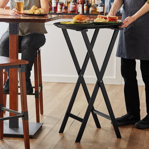 A woman standing at a black table with a Lancaster Table & Seating black wood folding tray stand holding food.