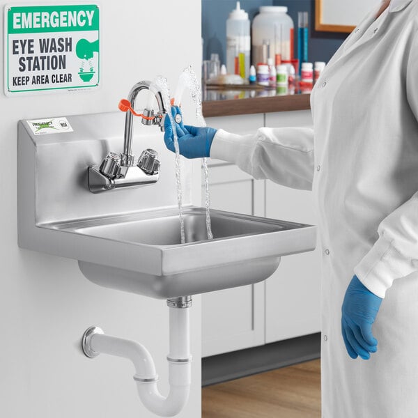 A woman in a blue lab coat and blue gloves washing her hands in a Regency wall mounted sink.