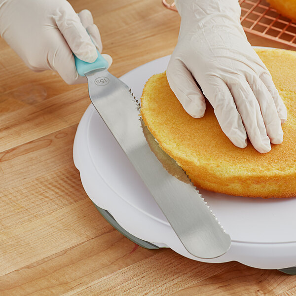 A hand in a glove using a Wilton straight serrated icing spatula to cut a cake.
