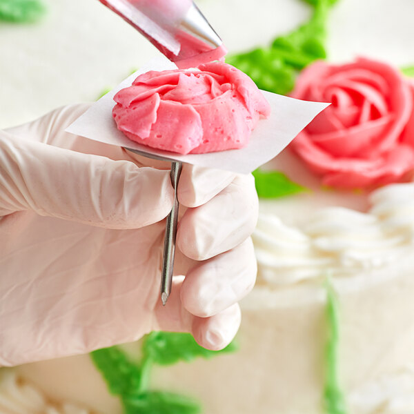 A person holding a Wilton stainless steel flower nail over a piece of cake with pink frosting.