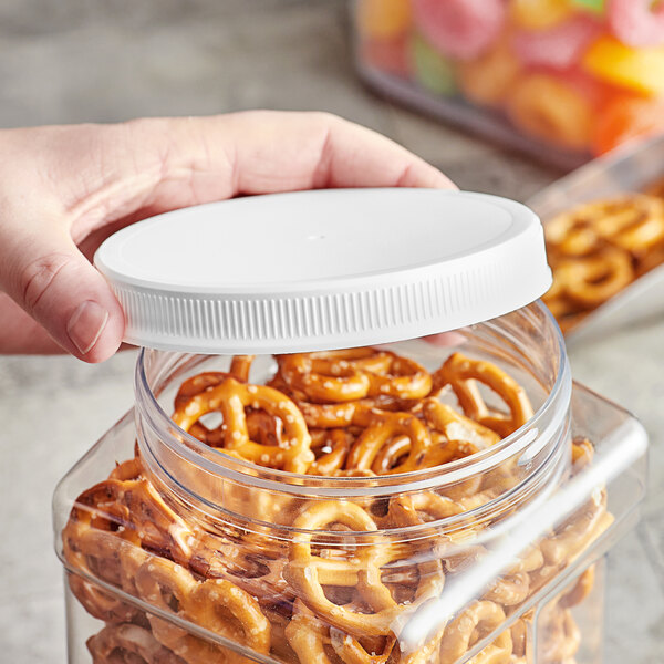 A hand holding a white plastic lid over a container of pretzels.