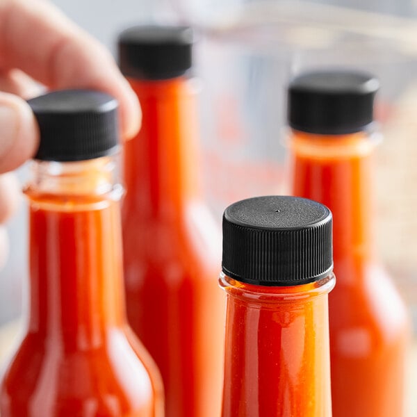 A hand holding a small bottle of red sauce with a black plastic lid.