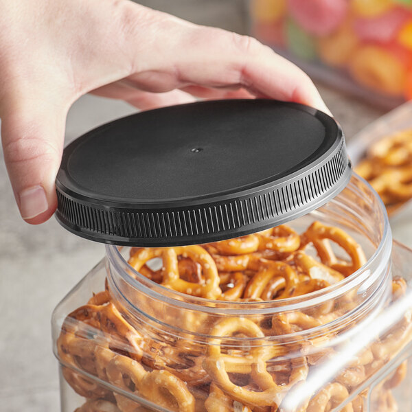 A hand holding a black plastic cap over a plastic container of pretzels.