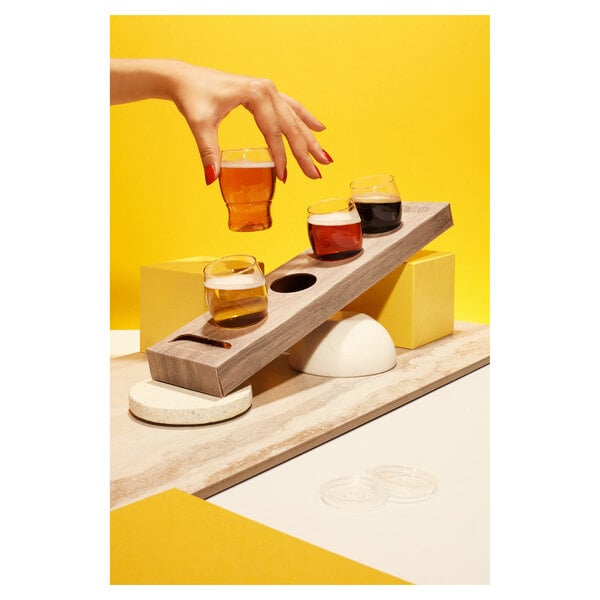 A hand holding a Tossware POP taster glass of beer on a wooden flight tray.