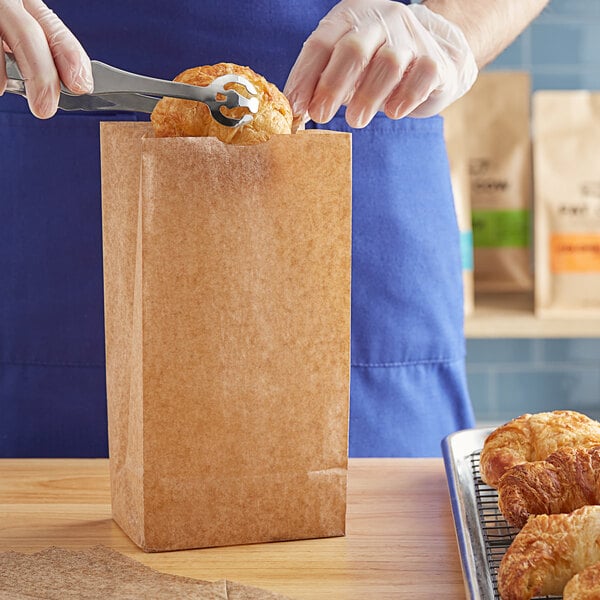 A person's gloved hands using scissors to open a Choice Natural Kraft Waxed Paper Bag containing a croissant.