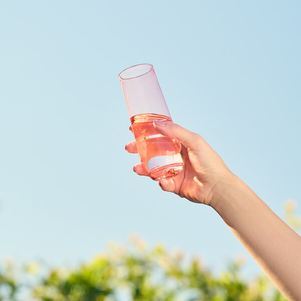 A hand holding a Tossware coral pink flute glass filled with pink liquid.
