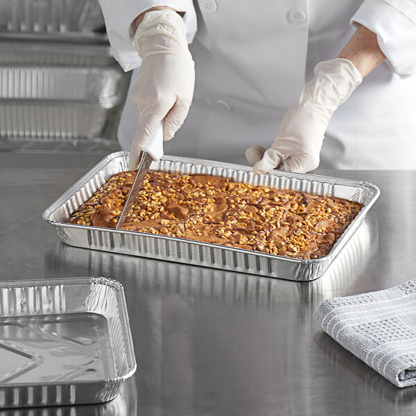 A person in gloves cutting a brown cake in a Baker's Lane foil cake pan.