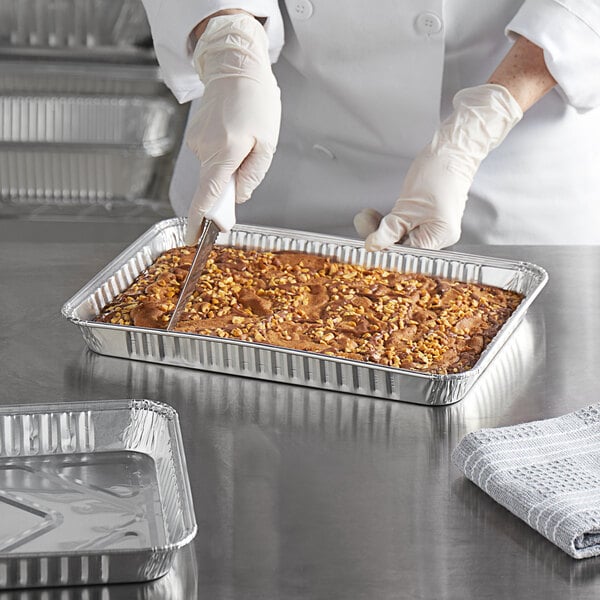 A person in gloves cutting a brown cake in a Baker's Mark foil cake pan.