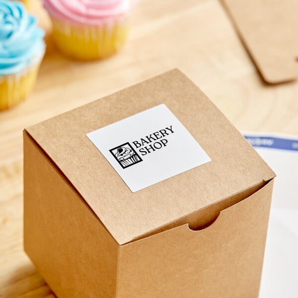 A brown box with a white label for Avery square labels on a counter with a cupcake.
