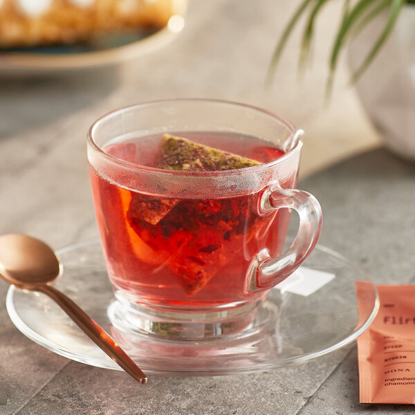 A glass cup of Dona Flirt herbal tea with a tea bag in it on a saucer with a spoon.