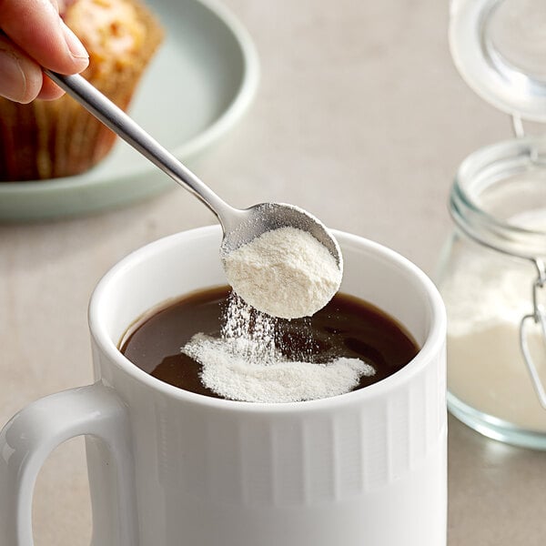 A hand holding a spoon scooping Nestle Coffee-Mate powder into a mug of coffee.