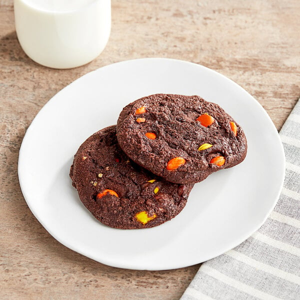 Two Otis Spunkmeyer chocolate cookies with REESE'S PIECES on a plate with a glass of milk.